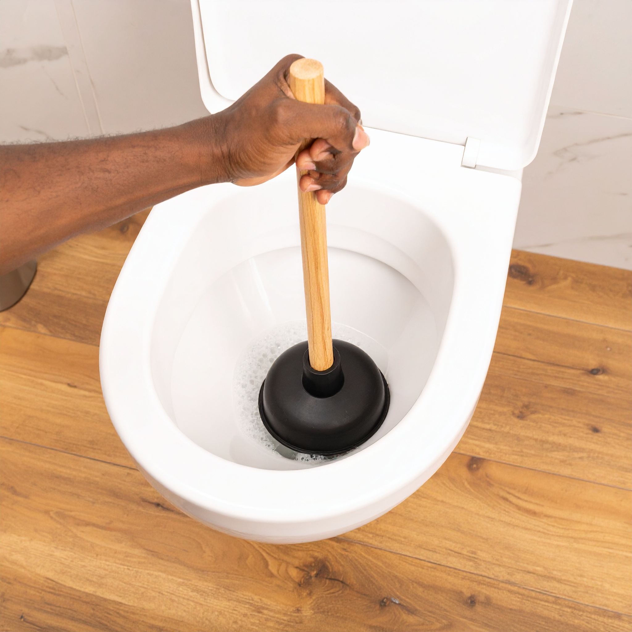 bathroom-toilet-clog Person using a plunger to unclog a toilet in a modern bathroom with wood flooring.